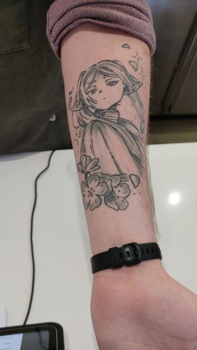 Freiren tatoo on a forearm. It shows the character with cherry blossom petals falling around here and stars in the upper right. Cherry blossom flowers are on the bottom left